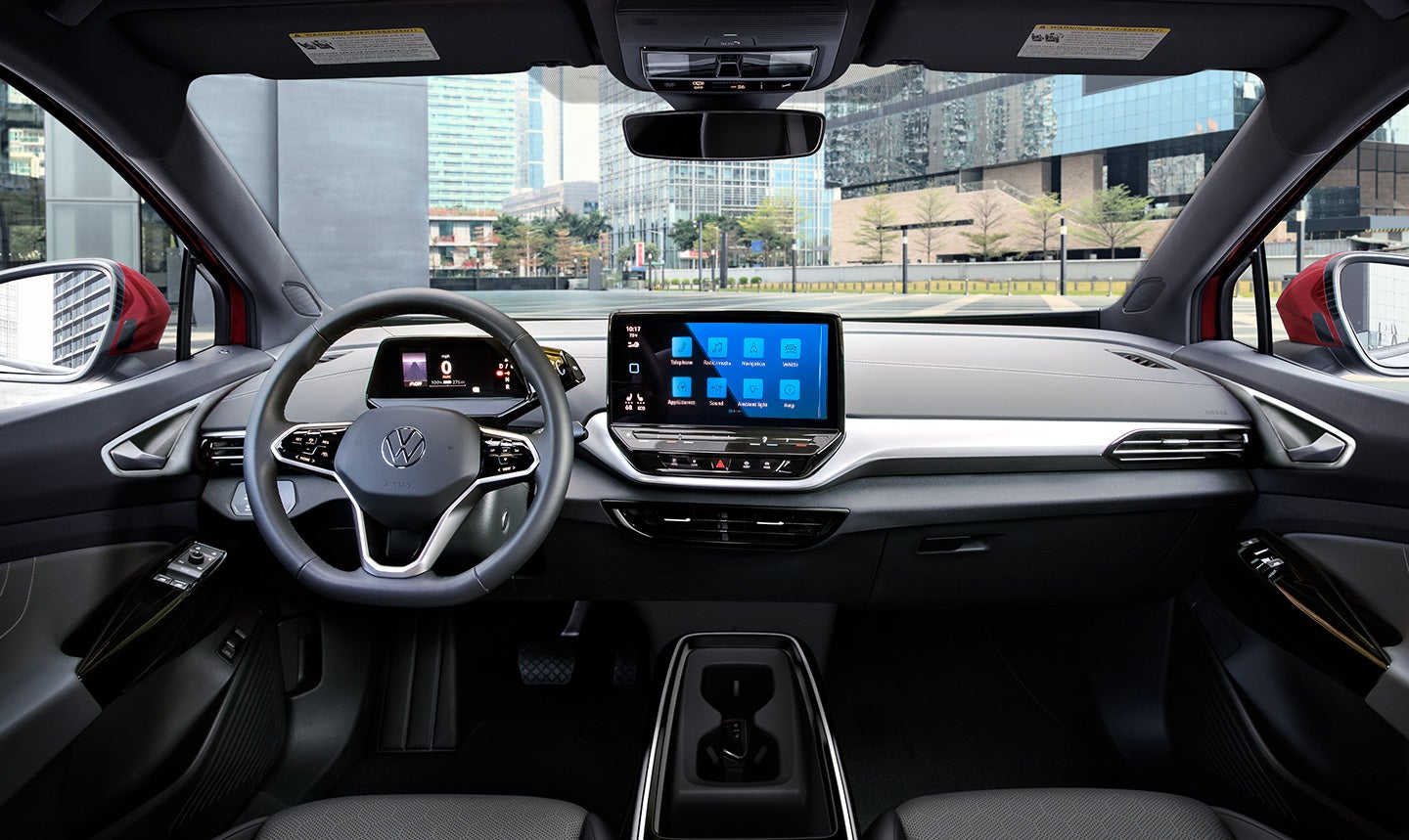 An interior shot of the ID.4 cockpit area featuring the Galaxy interior, steering wheel, driver display, and center console. Parked downtown, a modern city can be seen through the windshield.